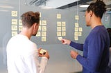 Two people, one wearing a long-sleeve white shirt, the other wearing a long-sleeve purple shirt, use post-it notes to plan the four quarters of a business.