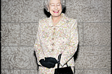 The late Queen Elizabeth the second standing in the center of the frame. She’s wearing a yellow floral print jacket and skirt. Wearing black gloves with a black purse hanging from her left wrist. Her eyes are closed and she’s smiling with lips painted red. It’s not a regular boring image of her because of the look on her face. Photograph by Christopher Wahl.