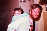 My dad and I, approximately 1984. Description: an older photograph of a white man with red hair and a longish beard with mustache and blue eyes looks at the camera and smiles. Me, a small white toddler of about 3, hangs on for a piggyback ride. I’m wearing a light blue shirt with long sleeves.