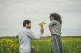 The picture shows a man and a woman in a beautiful green field. The man is down on one knee and has picked some yellow flowers from the field. He is holding them out to the woman. She is looking at him shyly. It’s unclear whether she will accept them or not.