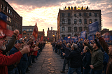 Two angry crowds face off in a cracked mill-town street at dusk while, unnoticed above them, suited figures dine at a glowing rooftop banquet table.