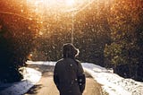 A person walks down a snowy path, surrounded by softly falling snow and warm sunlight filtering through the trees