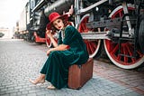 Woman in green dress with red hat sits on suitcase next to steam train