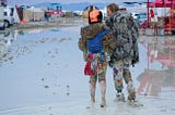 A couple seen from behind gaze at an expanse of flooded field while wading in mud.