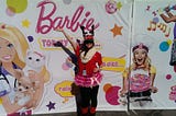 A girl in a red shirt with a reindeer hat, scarf, and tutu stands in front of a Barbie poster.