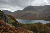 A rocky, tree-covered hill in the foreground, in front of a lake, with a mountain behind it. Grey cloudy skies above.