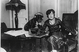 Black and white photo of Mme. Asta Souvrina, “the Bernhardt of Russia,” sitting beside a dog named Buster. Both are wearing headphones, listening to a radiophone device on a table in front of them. Taken around 1922 by Underwood & Underwood.