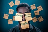 Homem com rosto coberto por post its com as palavras normas, expectativas, sociedade. Ao fundo, a parede também conta com mais post its com as palavras ódio, julgamento, dúvidas, gênero, sexualidade, racismo, fracasso, alma, entre outras.