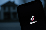 TikTok logo in pink-white-blue glow on smartphone screen at night, with blurred house in dark background.