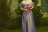 A redhead girl with her hair up wears a long, lavender, off-the-shoulder dress and holds a large red hibiscus flower in front of a huge bush in front of a ranch-style house.