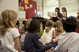 Children in a classroom being read to by a teacher.