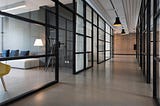 Image of office featuring hallway with glass doors leadign to meeting rooms. Meeting room on the left of the picture with couch and yellow chair.