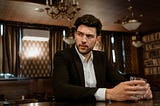 A man in a black suit with a drink, sitting at a bar in a sophisticated setting.