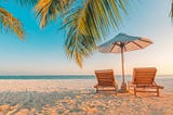 2 lounge chairs on a beach under a beach umbrella with palm leaves in the foreground