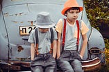 Two boys wearing gray shirts sitting on gray vehicle bumper