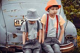 Two boys wearing gray shirts sitting on gray vehicle bumper