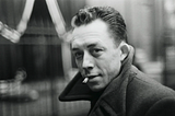 Black-and-white portrait of Albert Camus, a man in his thirties with combed-back dark hair, glancing over his shoulder toward the camera. He wears a heavy overcoat with the collar turned up and a lit cigarette between his lips, against a blurred, dimly lit background.