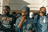A Black woman speaks at a rally with two Black men on either side of her. Several other Black people stand behind the trio.