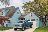 Picture of a cookie-cutter blue home in a lower middle class neighborhood with a pick up truck in front of it; the american dream encapsulatyed in an image.
