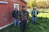 three people stand in front of a building with a wooden fence and trees in the background