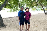 A woman and her daughter in scuba wear stand on the beach. There are green leaves on the trees on either side of them. This is a beach in a fertile place. The daughter is as tall as the mother.