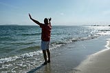 man standing at the sandy beach with the waves crashing into his feet and he rise his hand as if he embrance the morning sunny day and blue sky