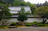 Green trees behind a rock garden, in a zen garden. In the background is a wall with Japanese style shingles.