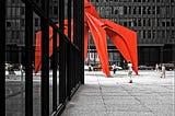 Half of a large red metal sculpture appears around the corner of a modern skyscraper, creating an illusion of symmetry, with that part of the red metal sculpture reflected in the building’s windows. A handful of relatively small people walk around in the middle distance.