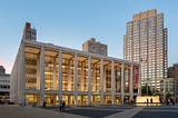 picture of a David Geffen Hall, a building with tall, thin rectangular columns marking the exterior of the building, which lets a viewer see the interior of the building through windows that extend from the ground to the roof. It is sunset and the windows are illuminated with gold light. A red New York Philharmonic banner hangs vertically between the two columns all the way to the far right.