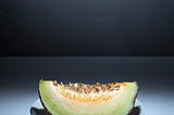 The image shows a single slice of cantaloupe melon placed on a white plate. The melon has a textured rind with a pale green and orange flesh, and its seeds are still attached. The background is plain and well-lit, emphasizing the fruit