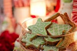 This is an image of Christmas cookies. It’s a close-up of a glass jar with star-shaped cookies with green and white icing. In the background, blurred, are red and white objects and a lamp, making for a nice overall Christmas-feel with red, white and green colours.