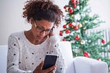 Black woman looking down at her phone with a concerned look on her face, with a Christmas tree in the background.