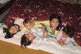 Five children, 4 sisters and a brother, laying down in a futon together. The three older children are smiling, the two younger children look ready to cry.