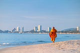 An orange-clad monk walking toward the camera on the beach, with sparse buildings on the horizon under a vast blue sky.