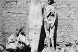 Black and white photo showing a man (on the left) sitting with his head in his hand, looking bored. Next to him, propped against a wall are Egyptian mummified corpses.