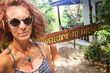 Author with big red hair, wearing shades and a spagetti-strapped dress in front of a sign that reads, “Welcome to High Bar.”