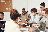 Group of colleagues collaborating around a table, with a Black man at the center, engaging in discussion with diverse team members.