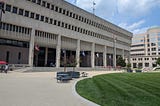 The front of the Towson Courthouse (Baltimore County Courts Building). A large light tan walkway is visible leading up to the steps, with a circular expanse of well-kept grass just to the front right. The building is a large modern building, light tan with columns and flags in the front.