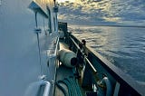 The starboard side of a ship can be seen. The ocean and a shoreline are in the photo