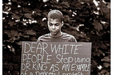 Young Black man holds a sign: “Dear White People: Stop using Dr. King as an example of peaceful protest…You Shot Him Too”