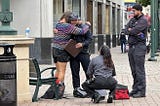 On a city sidewalk in Oakland, CA, a MACRO program responder kneels beside a person on the ground while another responder stands nearby and another person offers a supportive hug.