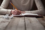 Writing with a pencil in a leather-bound notebook with crumpled drafts on a wooden table.