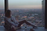 A midlife woman sitting alone beside a large apartment window overlooking a city skyline at dusk, relaxed in a calm modern space, reflecting quietly in peaceful solitude.