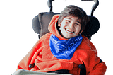 [Image: boy in comfy wheelchair and orange hoodie; he also has a blue bandana around his neck and a wire over his teeth.]