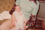 Newborn baby in sleeper with uncomfortable facial expression being held by adoptive mother with 70s style clothing and furninshings