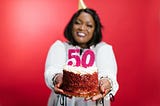 A Black woman with shoulder length straight hair and a party hat wears a white jumper with a pink number 50 on the front. She is smiling and holding a birthday cake, against a red backdrop