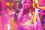 A double exposure depicting the author drinking a cocktail, staring into the camera, while another version of her takes a picture of the scene to the left. The image showing through from underneath her is a woman holding a beer, talking to someone outside the frame.