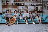 A family of six sit on a long couch on various internet enabled devices. They laugh at a dog on the floor.