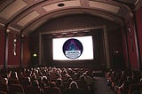 Folks in a movie theater staring at a screen that features the Dumpster Fire logo of While Rome Burns.