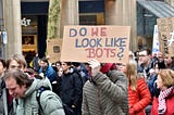 People walking at the demostration, one guy is holding a sign “DO WE LOOK LIKE BOTS?”